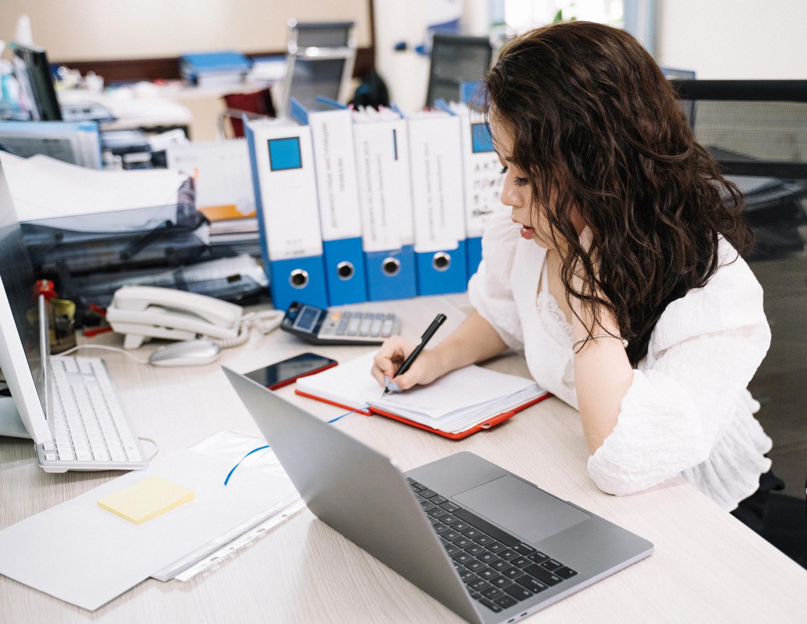 Eine Frau schreibt an einem vollen Büroschreibtisch mit Ordnern, Laptop und Telefon in ein Notizbuch.