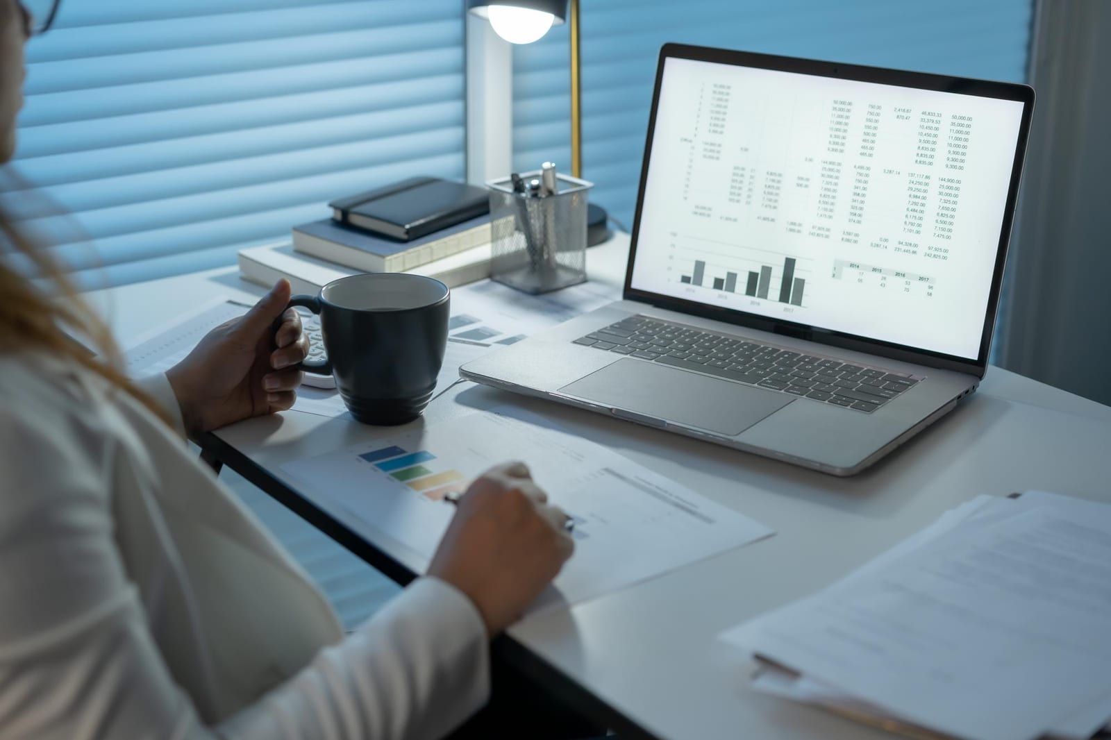Eine Frau am Schreibtisch mit einem Laptop, auf dem eine Tabelle geöffnet ist, dazu eine Tasse und gedruckte Diagramme am Abend.